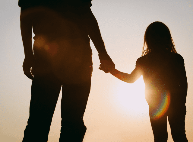 Silhouette of an adult holding a child's hand with the sunset in the distance.